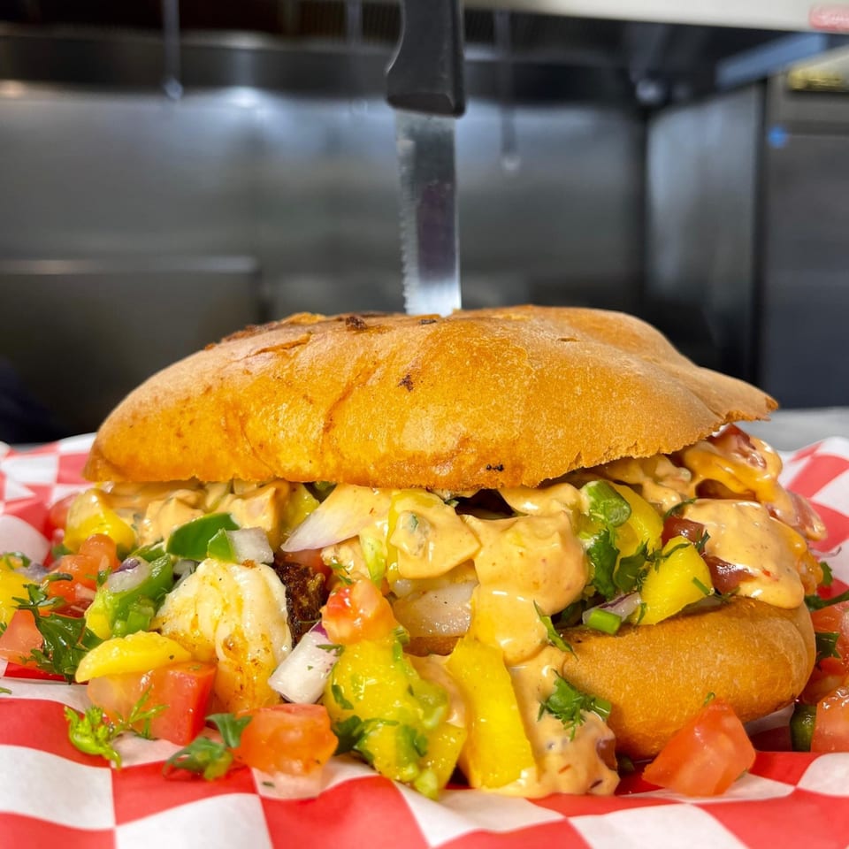A large Mexican torta sandwich filled with shrimp, mango, and a creamy sauce, served on a red and white checkered paper.
