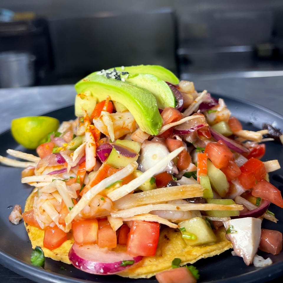 A vibrant Tacos Acambaro tostada piled high with seafood, diced tomatoes, onions, and avocado slices.