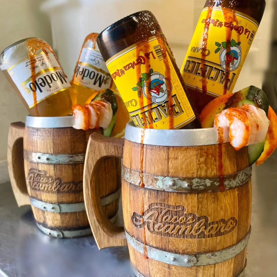 Two Tacos Acambaro wooden mugs with Modelo and Pacifico beers, shrimp, and cucumber garnishes.